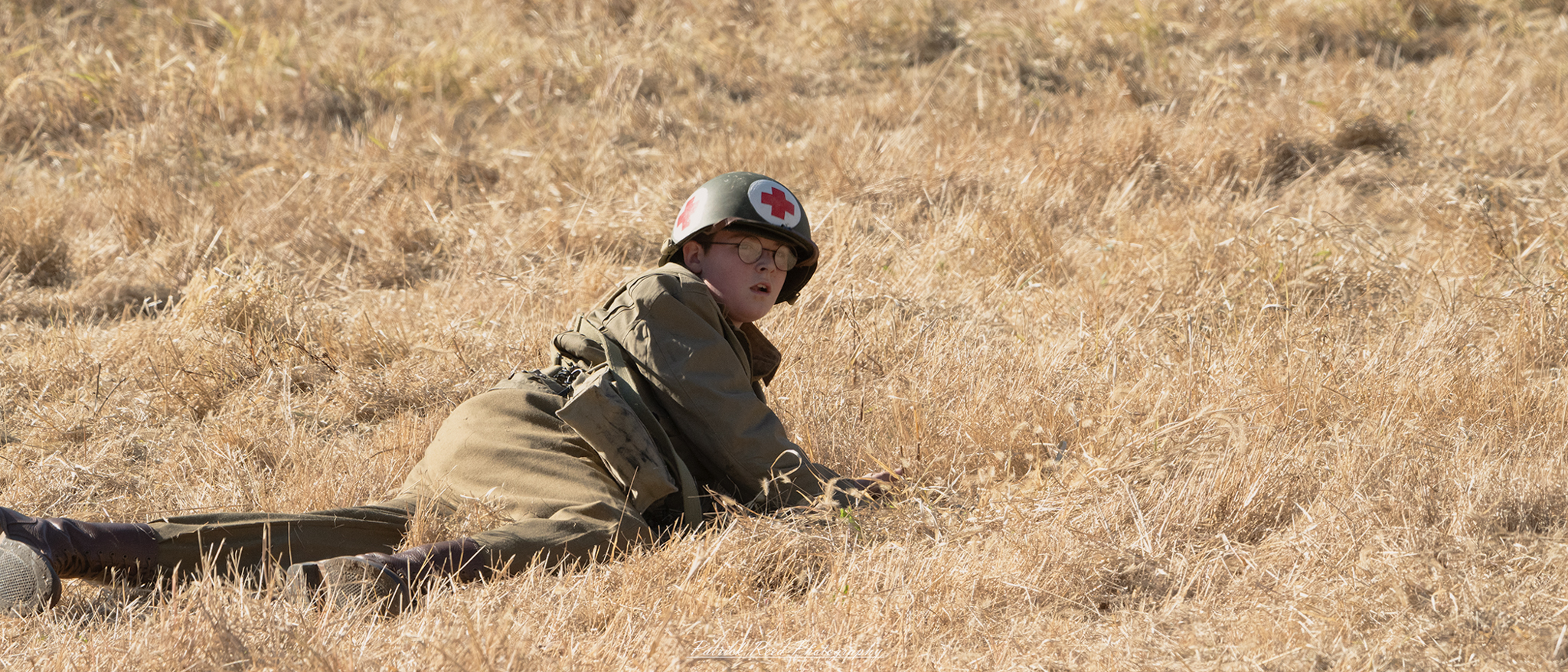 A young WWII medic lies in a grassy field, surrounded by the remnants of battle. He is dressed in a worn uniform, with a distinctive red cross armband. His face shows signs of exhaustion and concern as he stares into the distance, perhaps reflecting on the weight of his responsibilities. The field is dotted with wildflowers and hints of conflict, creating a stark contrast to the peaceful surroundings. The scene captures the solitude and challenges faced by medics in wartime, highlighting their bravery and dedication to saving lives amidst chaos.